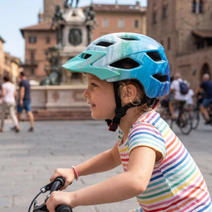 A little girl wearing an ocean blue helmet is riding a bicycle in the square